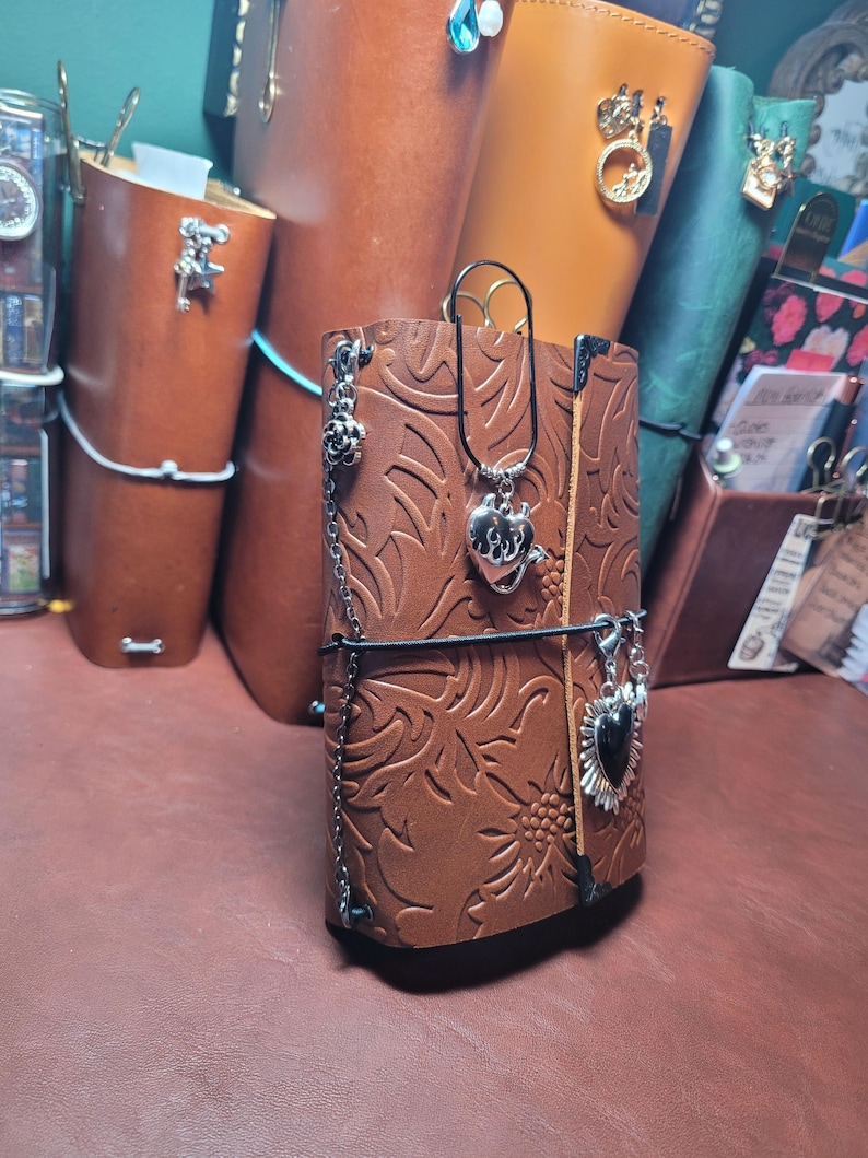 May include: A brown leather journal with embossed floral patterns. The journal is adorned with silver charms and a black elastic closure. Other leather journals in various colors are visible in the background.