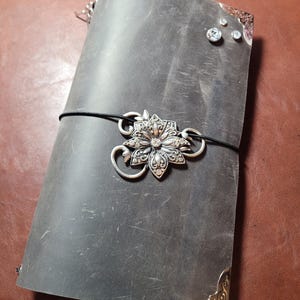 May include: A dark gray leather journal with a decorative silver flower clasp and corner accents. The journal is secured with a black elastic band. Rhinestones are affixed to the cover.