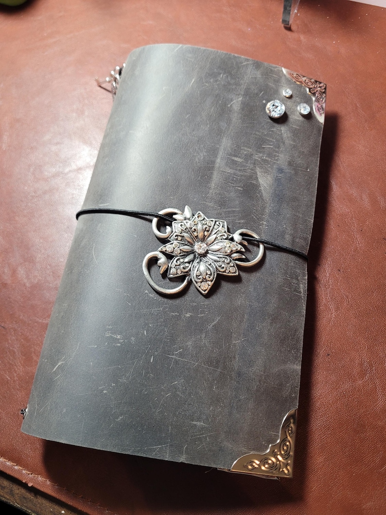 May include: A dark gray leather journal with a decorative silver flower clasp and corner accents. The journal is secured with a black elastic band. Rhinestones are affixed to the cover.