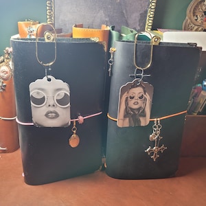 May include: Two black leather journals with decorative charms and photo pendants. One journal has a pink elastic band and a gold charm. The other has an orange band and a silver cross charm. Both have gold paperclip-style clasps.