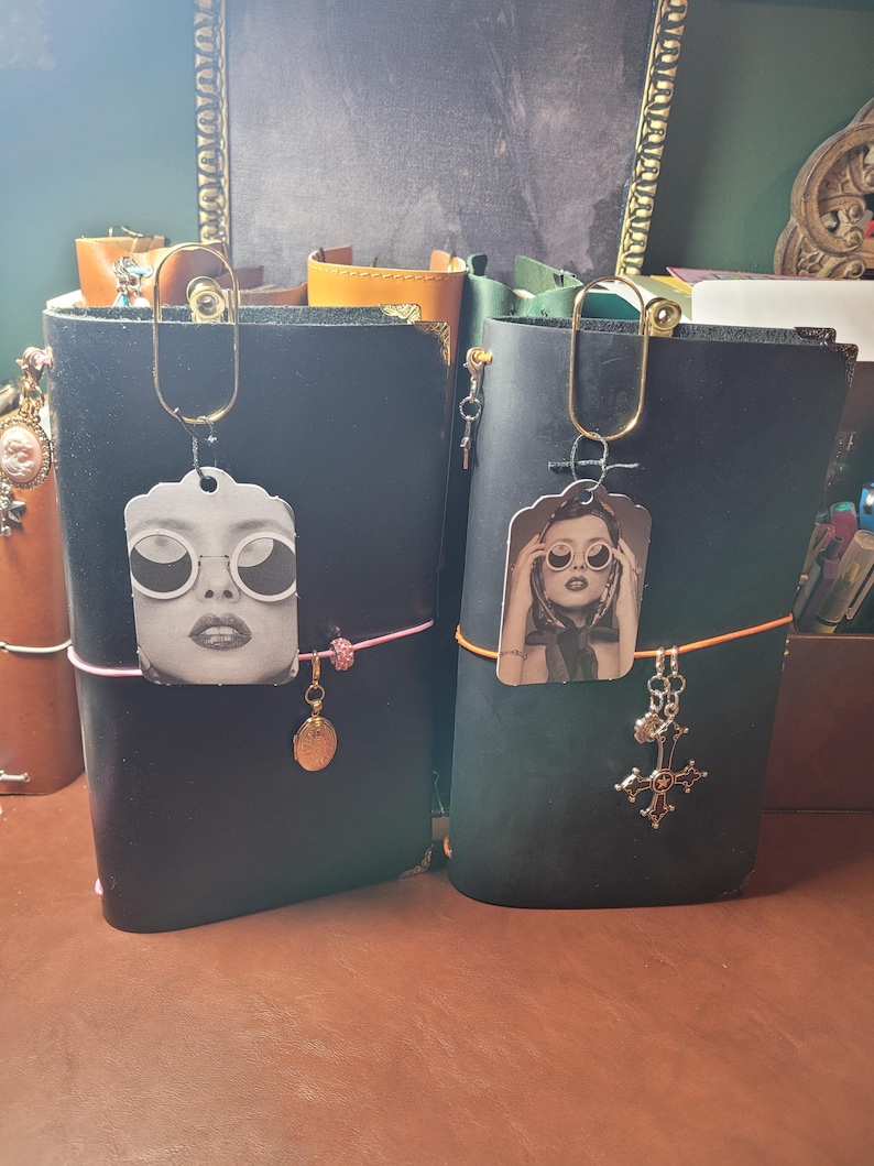 May include: Two black leather journals with decorative charms and photo pendants. One journal has a pink elastic band and a gold charm. The other has an orange band and a silver cross charm. Both have gold paperclip-style clasps.