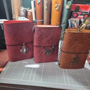 May include: Three leather journals in shades of red and brown, featuring embossed floral patterns. Each journal has a decorative charm and a black elastic closure. The journals are displayed on a gray cutting mat.