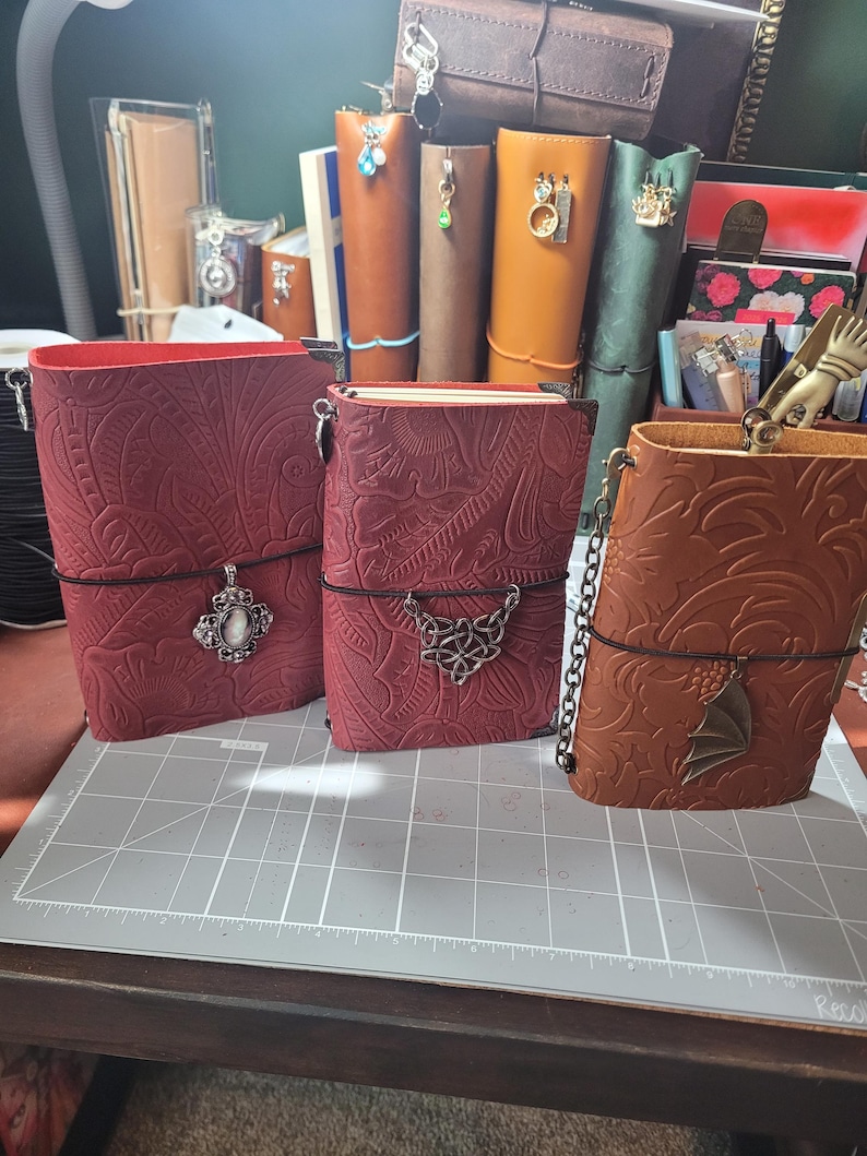 May include: Three leather journals in shades of red and brown, featuring embossed floral patterns. Each journal has a decorative charm and a black elastic closure. The journals are displayed on a gray cutting mat.