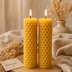 Può includere: Due candele gialle accese con motivo a nido d'ape su una fetta di legno. Visibile un biglietto con la scritta "Grazie" e un cuore. Grano secco e un vaso sullo sfondo. Arredamento domestico caldo e invitante.