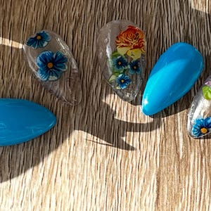 Peut inclure: Plusieurs faux ongles aux motifs variés. Certains sont turquoise uni, d'autres présentent des motifs floraux avec des fleurs bleues et jaunes. Un ongle arbore une rose orange avec des feuilles vertes.