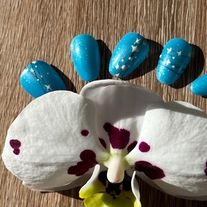 Peut inclure: Une fleur d'orchidée blanche avec des taches violettes et un centre jaune est présentée avec plusieurs ongles bleus. Les ongles ont une finition scintillante et des motifs d'étoiles blanches. Le fond est une surface en bois.