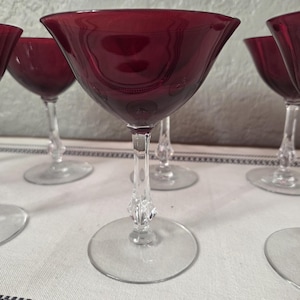 May include: A set of ruby red wine glasses with a flared bowl design. The glasses have clear, ornate stems and circular bases. The glasses are arranged on a white tablecloth with a black and white patterned border.