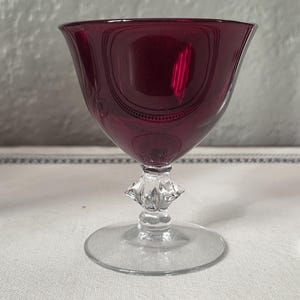 May include: A ruby-colored glass goblet with a clear, faceted stem and a round base. The bowl of the glass has a decorative etched design. The glass is set on a white tablecloth with a black and white border.