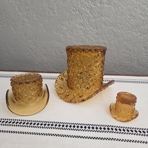 May include: Three amber-colored glass top hat-shaped containers. The largest is a top hat, the second is a cowboy hat, and the smallest is a top hat. All have a diamond pattern. They are displayed on a white and black patterned surface.