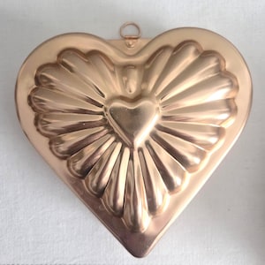 May include: A copper-colored heart-shaped mold with a central heart design and radiating lines. The mold has a loop at the top for hanging. The vintage kitchenware is likely used for baking or decorative purposes.