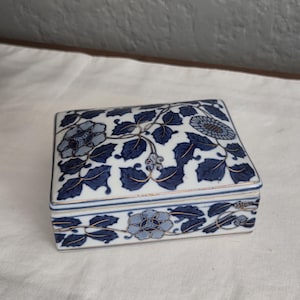 May include: A rectangular porcelain trinket box featuring a white base with a blue floral pattern and gold-toned trim. The lid and sides are decorated with detailed blue leaves and flowers, showcasing a consistent color palette throughout the piece.