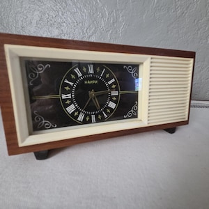 May include: Vintage rectangular clock with a dark face, Roman numerals, and gold accents. The clock is encased in a wooden frame with a cream-colored side panel. The clock has a retro design.