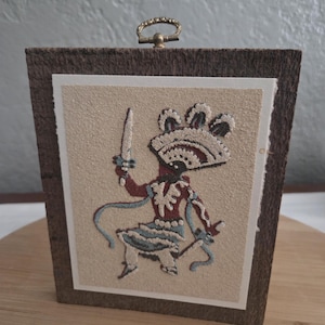 May include: A framed artwork featuring a stylized figure in a colorful costume holding a blade. The artwork is set against a textured beige background, framed in dark brown, and has a small brass handle at the top.