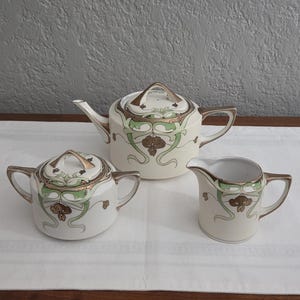May include: A cream-colored tea set with gold and green Art Nouveau-style floral designs. The set includes a teapot, a sugar bowl with a lid, and a creamer. The set is displayed on a white cloth.