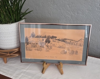 May include: A framed landscape drawing on tan paper featuring a rural scene with a small house, a barn, trees, and a fence. The artwork is enclosed in a thin metallic frame with a dark gray mat and includes the handwritten text "Camp at Malheur Dam".
