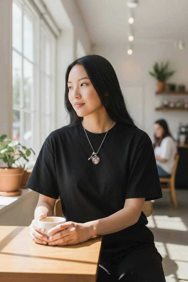 May include: A woman wearing a black t-shirt and a silver necklace with a pendant. The pendant has a heart-shaped design with red and white accents. She is holding a white cup of coffee and sitting at a wooden table near a window.