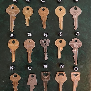 Puede incluir: Una colección de quince llaves antiguas, cada una con un diseño y forma únicos. Las llaves están dispuestas en un patrón de cuadrícula sobre una superficie verde oscuro, con cuentas negras con letras encima de cada llave. Las llaves son de latón.