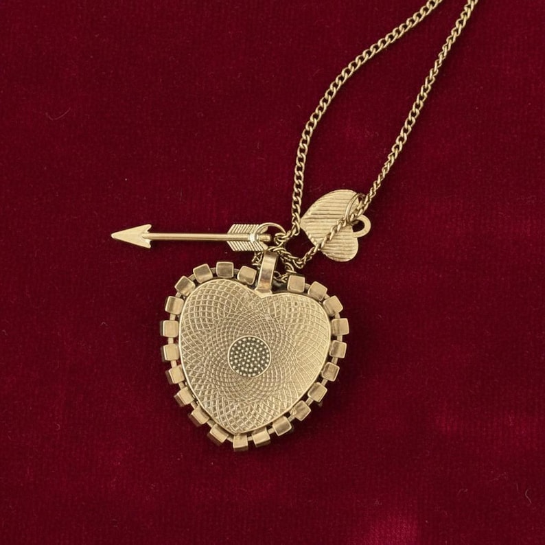 May include: A gold-colored heart-shaped pendant necklace with an arrow and a small heart charm. The heart pendant has a textured surface and a small circular design in the center. The necklace is on a gold chain.
