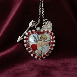 May include: Silver-toned necklace featuring a heart-shaped pendant with a vintage illustration of Cupid. The pendant is framed with red and clear crystals. An arrow charm and a wing charm are also attached to the chain.