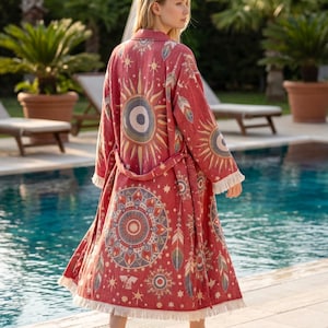 Peut inclure: Un long peignoir rouge ouvert sur le devant avec une ceinture à nouer et une bordure à franges à l'ourlet. Le tissu présente un motif répétitif de grands mandalas circulaires, de soleils et de plumes dans des tons de blanc, bleu et or.