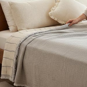 May include: A light grey bedspread with a fringed edge covers a bed with cream-coloured pillows and a wooden headboard. A person sits on the bed, wearing a beige pyjama set, and holding a magazine.