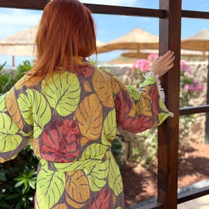May include: A colorful leaf-patterned robe with a matching belt. The robe features a vibrant design of green, orange, and red leaves on a gray background. The sleeves have a white fringe trim. The person is standing near a wooden structure, with a view of the ocean and beach umbrellas.