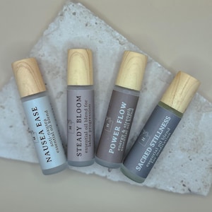 Puede incluir: Cuatro mezclas de aceites esenciales en botellas roll-on con tapas de madera. Las botellas están etiquetadas como "Nausea Ease", "Steady Bloom", "Power Flow" y "Sacred Stillness". Las botellas están dispuestas sobre una superficie de piedra blanca.