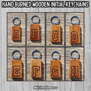 May include: Hand-burned wooden initial keychains. Each keychain features a rectangular wooden tag with a unique initial from A to H, burned in a bold, black font. The keychains have a silver metal ring and a blue loop.