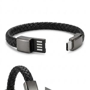Puede incluir: Pulsera de cuero trenzado negro con un conector USB y un conector USB-C. La pulsera tiene cierres y conectores de color plomo. El conector USB tiene cuatro líneas verticales.