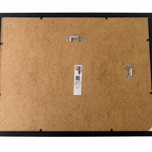 May include: The back of a rectangular black picture frame featuring a brown fiberboard backing. Two silver metal hanging brackets are attached to the board, one centered at the top and one on the right side. A white product label is centered.