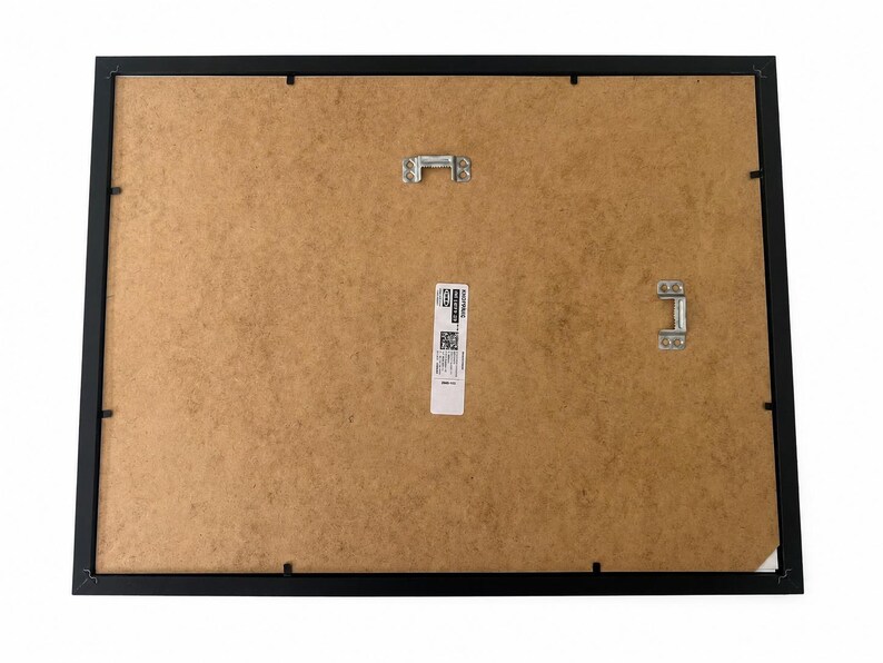 May include: The back of a rectangular black picture frame featuring a brown fiberboard backing. Two silver metal hanging brackets are attached to the board, one centered at the top and one on the right side. A white product label is centered.