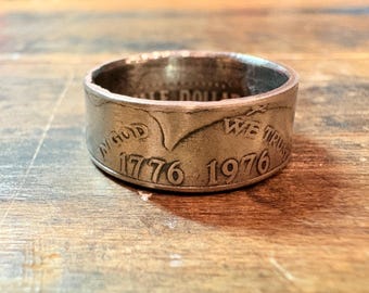May include: A silver ring made from a half dollar coin. The ring features the inscriptions "IN GOD WE TRUST", "1776", and "1976". The ring is a unique piece of jewelry, showcasing a vintage coin design.
