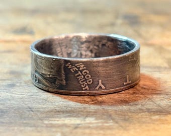 May include: A silver ring with the words "IN GOD WE TRUST" engraved on the side. The ring appears to be made from a coin, with details of the original design visible. The ring is set against a wooden surface.