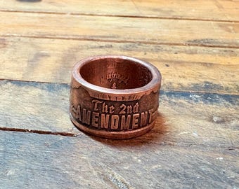 May include: A copper-colored ring with the text "The 2nd AMENDMENT" engraved around the band. The ring has a rustic, worn appearance, suggesting it is handmade or vintage. The inner band has additional text.