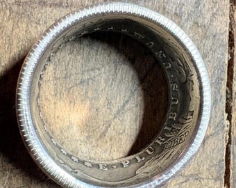 May include: A silver ring crafted from a coin, featuring intricate details of the original coin's design. The ring has a textured outer edge and visible inscriptions, showcasing the coin's history and craftsmanship. The ring is circular.