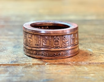 May include: A copper-toned metal ring featuring a series of embossed rectangular symbols around the band. The lower portion of the ring displays the engraved date 12-21-2012. The band has a flat, wide profile with a metallic finish and visible texture.