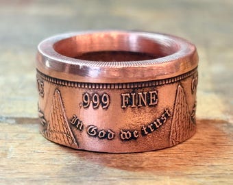 May include: A copper ring crafted from a coin, featuring a smooth, polished top edge and a textured outer band. The exterior displays the embossed text .999 FINE and In God we trust, alongside engraved triangular mountain-like shapes.