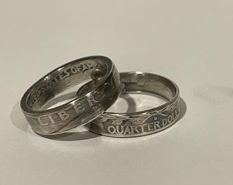 May include: Two silver rings made from US quarter coins. One ring displays the word "LIBERTY", and the other ring shows the words "QUARTER DOLL". The rings are on a white surface.