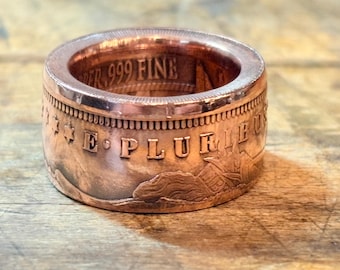 May include: A copper ring crafted from a coin, featuring the embossed text E PLURIBUS and stars on the outer band. The inner surface displays the text 999 FINE COPPER. The metal has a polished, reddish-brown metallic finish with visible relief details.