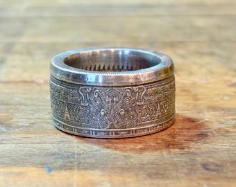 May include: A silver-toned coin ring featuring a wide band with intricate, embossed patterns of stylized figures and geometric motifs. The exterior surface displays a detailed relief design, while the interior edge shows a serrated coin-like texture.
