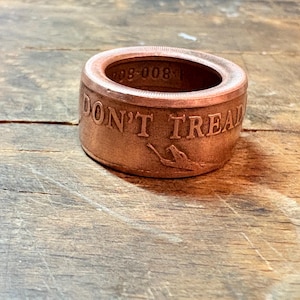 May include: A copper-colored ring with the words "DON'T TREAD" engraved on the side, along with a snake illustration. The ring has a circular design and is set against a wooden background.