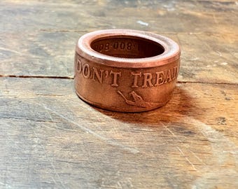 May include: A copper-colored ring with the words "DON'T TREAD" engraved on the side, along with a snake illustration. The ring has a wide band and a circular opening. The ring is sitting on a wooden surface.