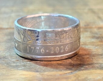May include: A silver-toned metal ring crafted from a coin, featuring a wide band with a textured surface. The exterior displays the engraved text 1776-2026, accompanied by small star symbols and intricate relief details visible along the upper edge.