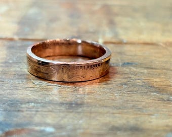 May include: A copper-colored ring crafted from a coin, featuring a smooth band with visible metallic texture. The exterior surface displays partial embossed lettering that reads URIBUS, along with a faint arrow-like design element.
