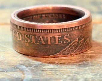 May include: A copper-colored ring with the words "UNITED STATES" embossed on the side. The ring has a detailed design and is set against a wooden surface. The ring appears to be made from a coin.