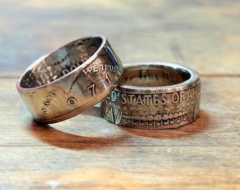 May include: Two silver coin rings are displayed on a wooden surface. One ring is tilted, revealing the inscription "IN GOD WE TRUST" and the number "7". The other ring shows "UNITED STATES OF AMERICA".