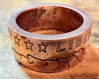 May include: A copper-colored ring with the word "LIBE" and three stars engraved on the band. The ring has a textured surface and appears to be made from a coin. The ring is approximately 1 inch wide.