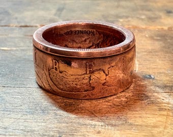 May include: A copper-colored ring with a wide band. The ring appears to be made from a coin, with visible details of the original design. The ring sits on a wooden surface.