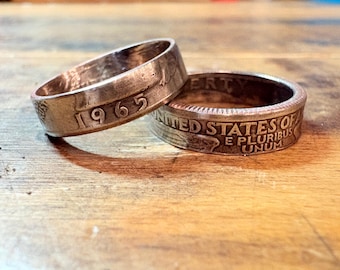 May include: Two handcrafted rings made from coins. One ring displays the year "1965", while the other features the text "UNITED STATES OF AMERICA" and "E PLURIBUS UNUM". The rings are made of a silver-colored metal and rest on a wooden surface.