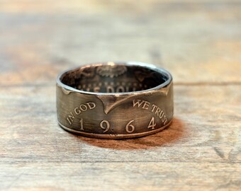 May include: A silver ring made from a 1964 coin. The ring features the inscriptions "IN GOD WE TRUST" and "1964" on its outer band. The ring is set against a wooden surface.